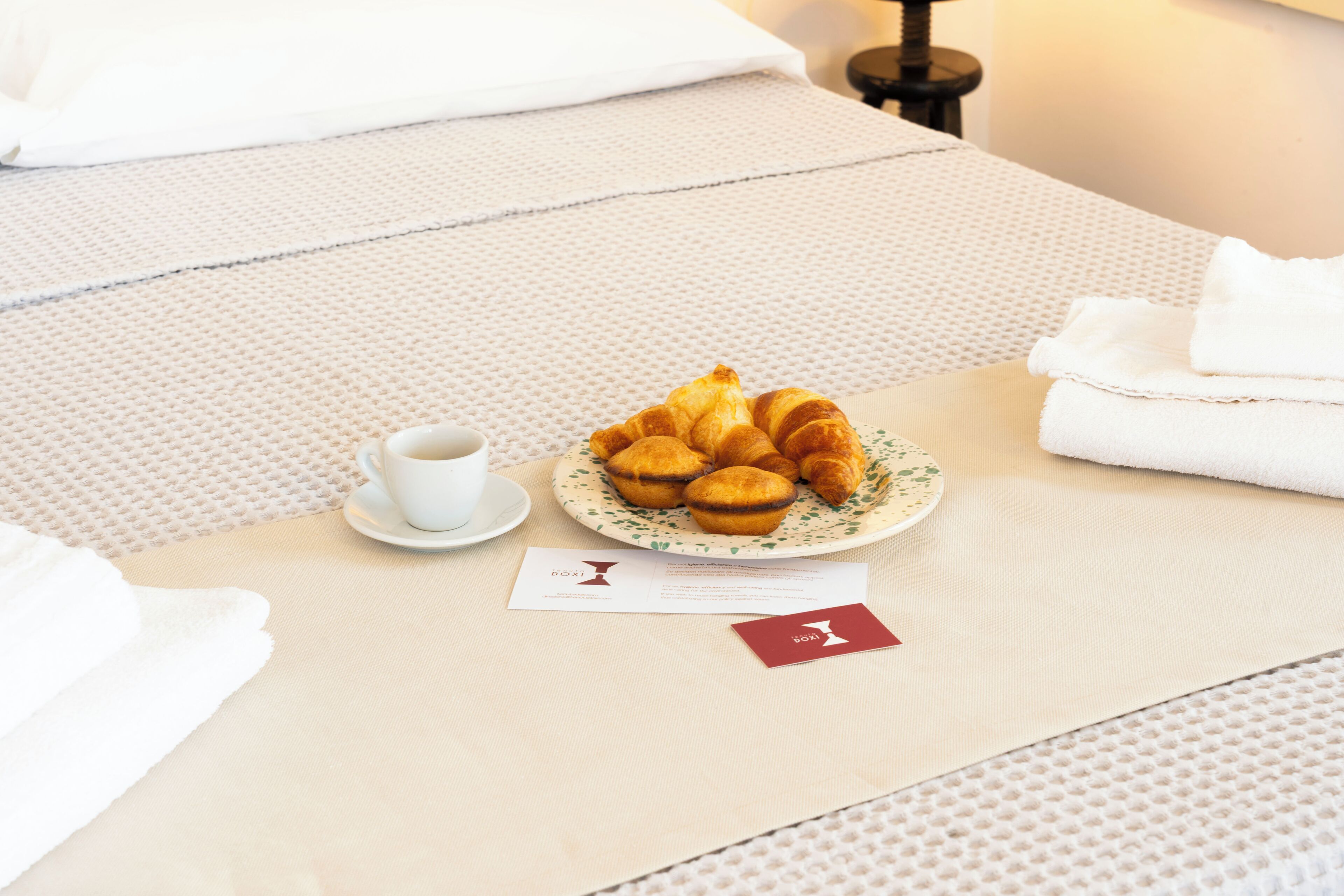 Una cama bien hecha con un plato de cruasanes, una taza de café y una tarjeta de presentación sobre una colcha con textura.