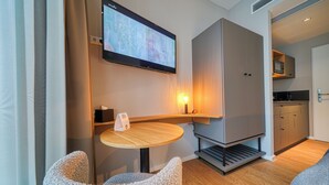 Ein modernes Hotelzimmer mit flachbildfernseher an der Wand, einem Holztisch mit Stuhl, einer eingebauten Sitzbank und einer Küchenzeile.