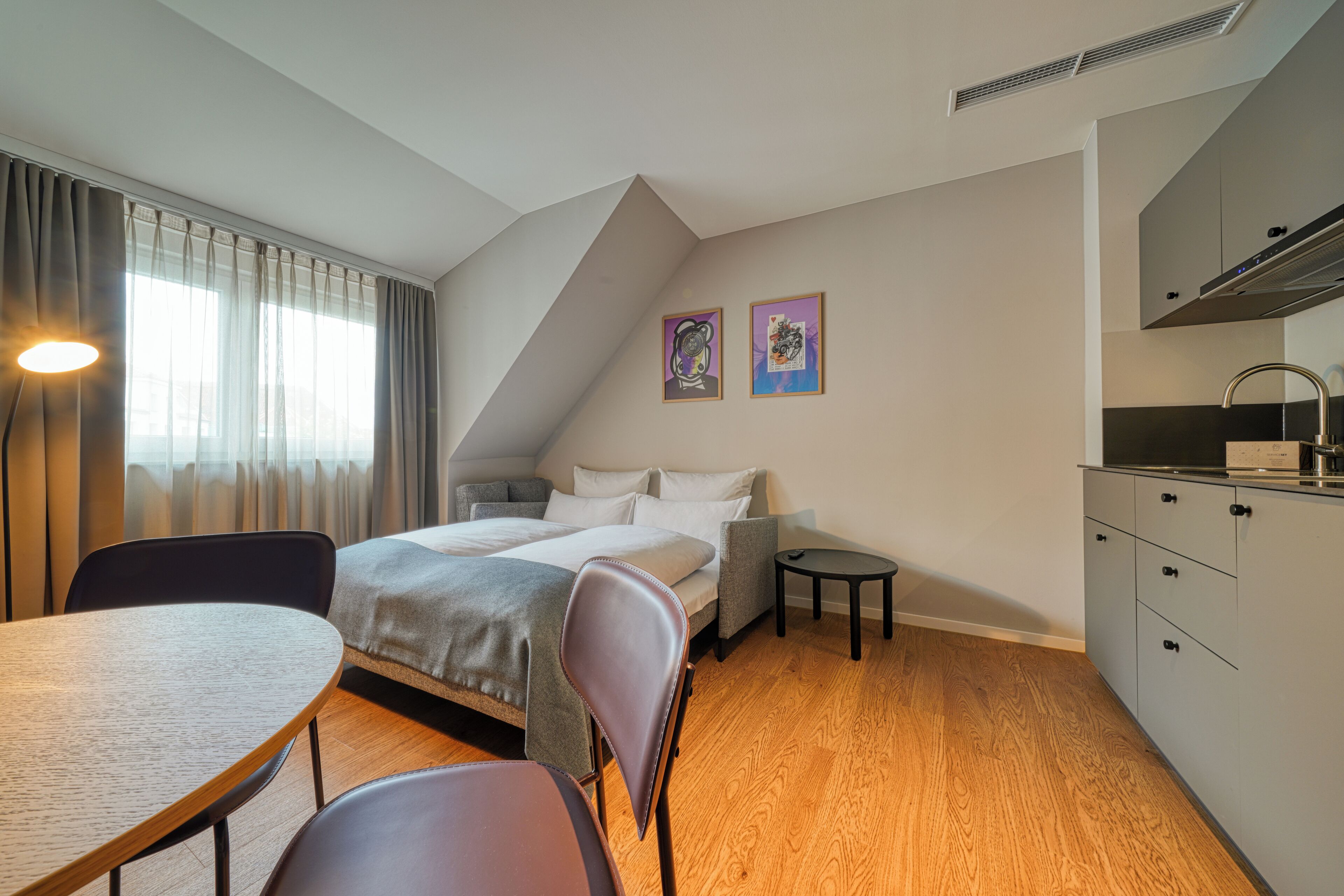 Habitación de hotel compacta con cama, pequeña mesa de comedor, kitchenette y ventana con cortinas.