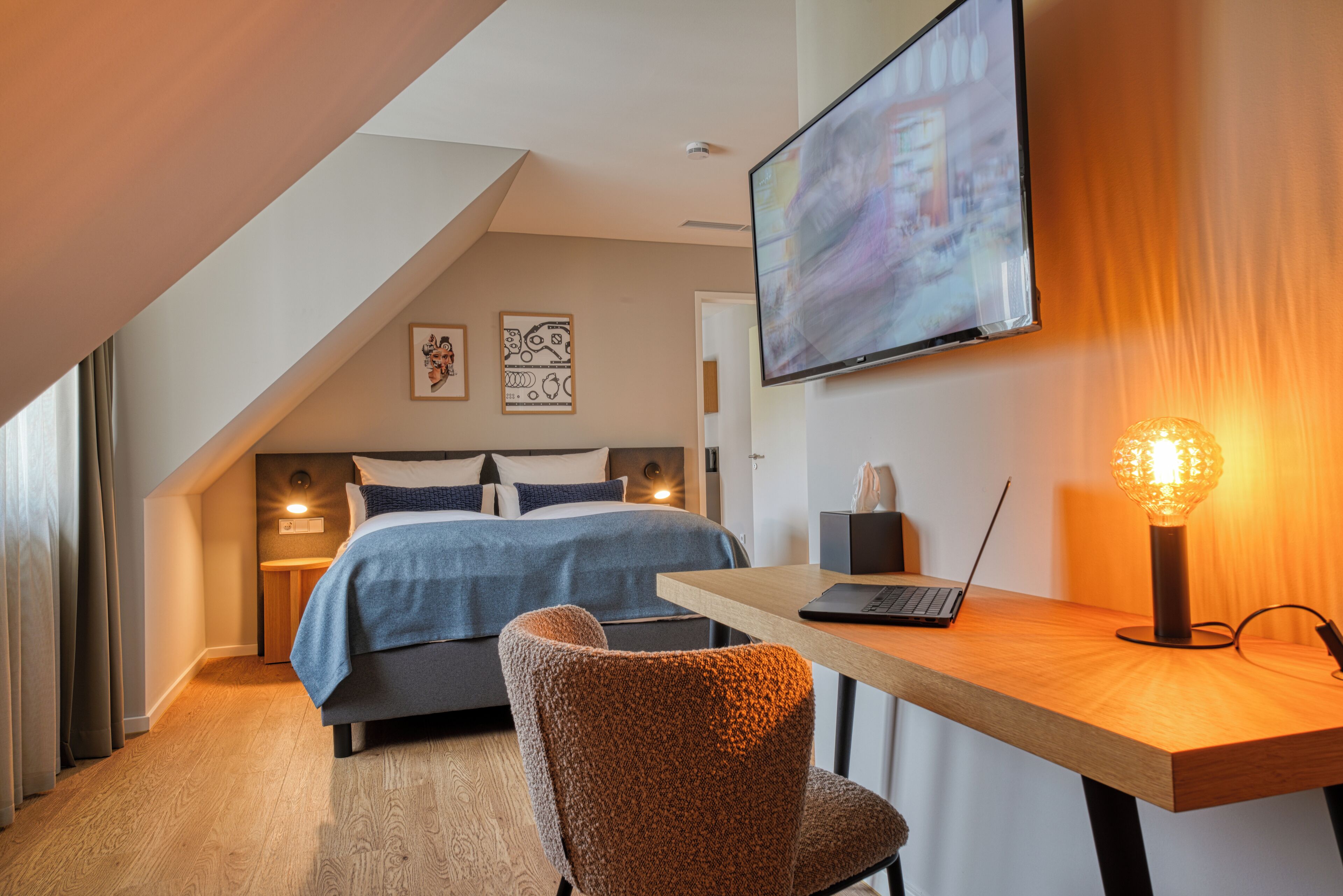 Quarto moderno com uma cama, uma escrivaninha com um laptop, uma luminária e uma TV na parede.