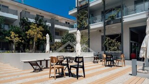 Een terras buiten met houten tafels en stoelen, omringd door een modern gebouw met balkons en beplanting.