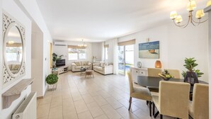 A spacious living area with a sofa set, a dining table with chairs, a TV, and a large window. - SunnyVillas: 3BR Family Villa☆Private Pool (Protaras)