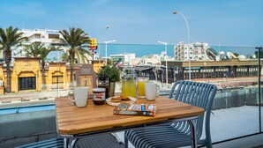 A table with a book, a Nutella jar, a glass of juice, and a coffee cup on a balcony overlooking a cityscape with palm trees and buildings.
