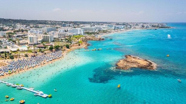 An aerial view of a coastal city with a beach, clear blue waters, and a rocky outcrop.