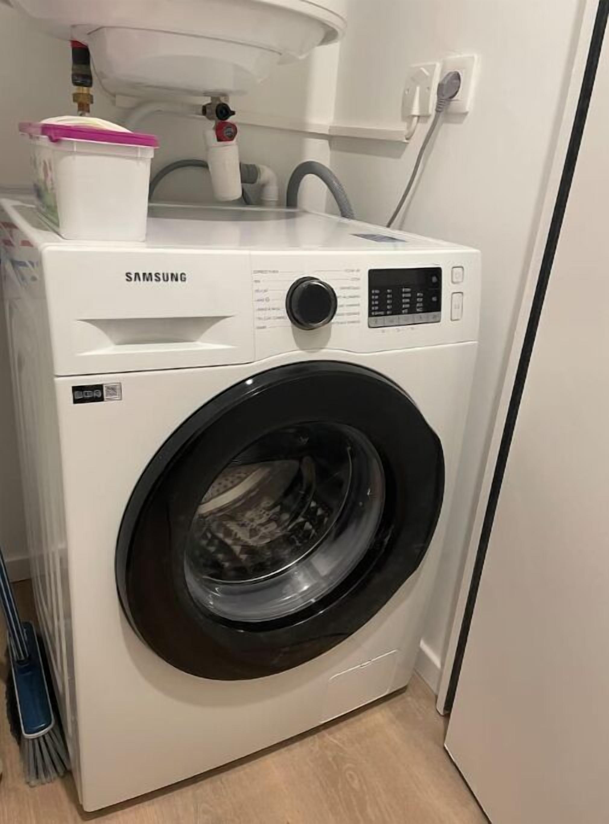 A Samsung front-loading washing machine in a laundry room with a white sink and a faucet above it.
