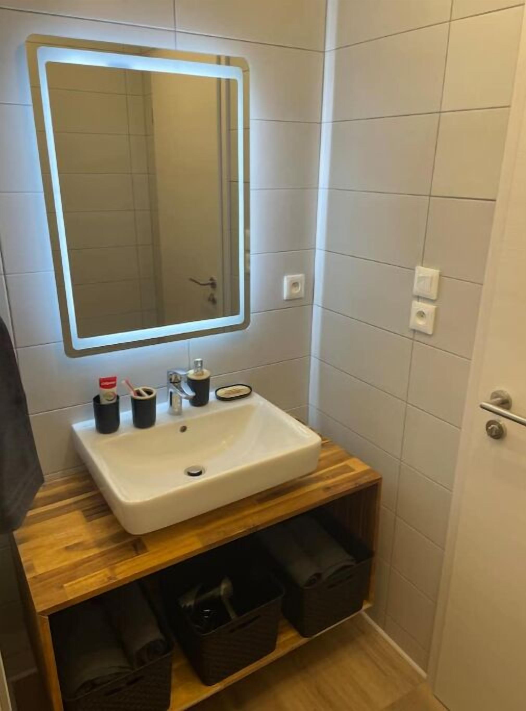 Een badkamer met een wit wastafel, een spiegel met verlichting, en twee zwarte vuilnisbakken.