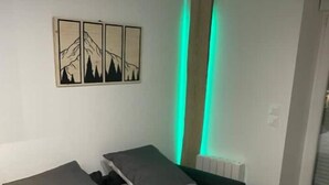 A single bed with white bedding and two pillows, a small bedside table, and a wall-mounted artwork of mountain silhouettes. - Appartement - Au Cœur du Lac Blanc (Orbey)