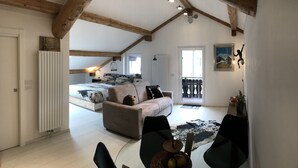Un vaste espace de vie doté d’un haut plafond, de poutres en bois, d’un canapé, d’une table à manger avec des chaises et d’un lit agrémenté d’un tableau représentant une montagne.