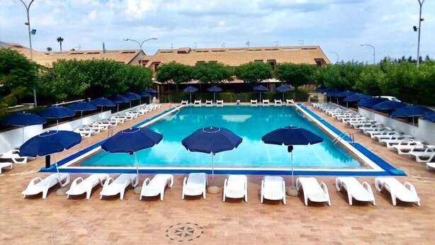 A pool area with sun loungers and umbrellas, surrounded by greenery and a building in the background. - Riviera Village (Butera)