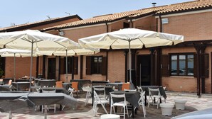 An outdoor seating area with tables and chairs, covered by umbrellas, in front of a brick building with windows and a tiled roof. - Riviera Village (Butera)