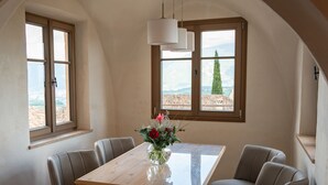 Un'area pranzo con un tavolo e sedie in legno, un vaso con fiori e ampie finestre che offrono vista su edifici e montagne.