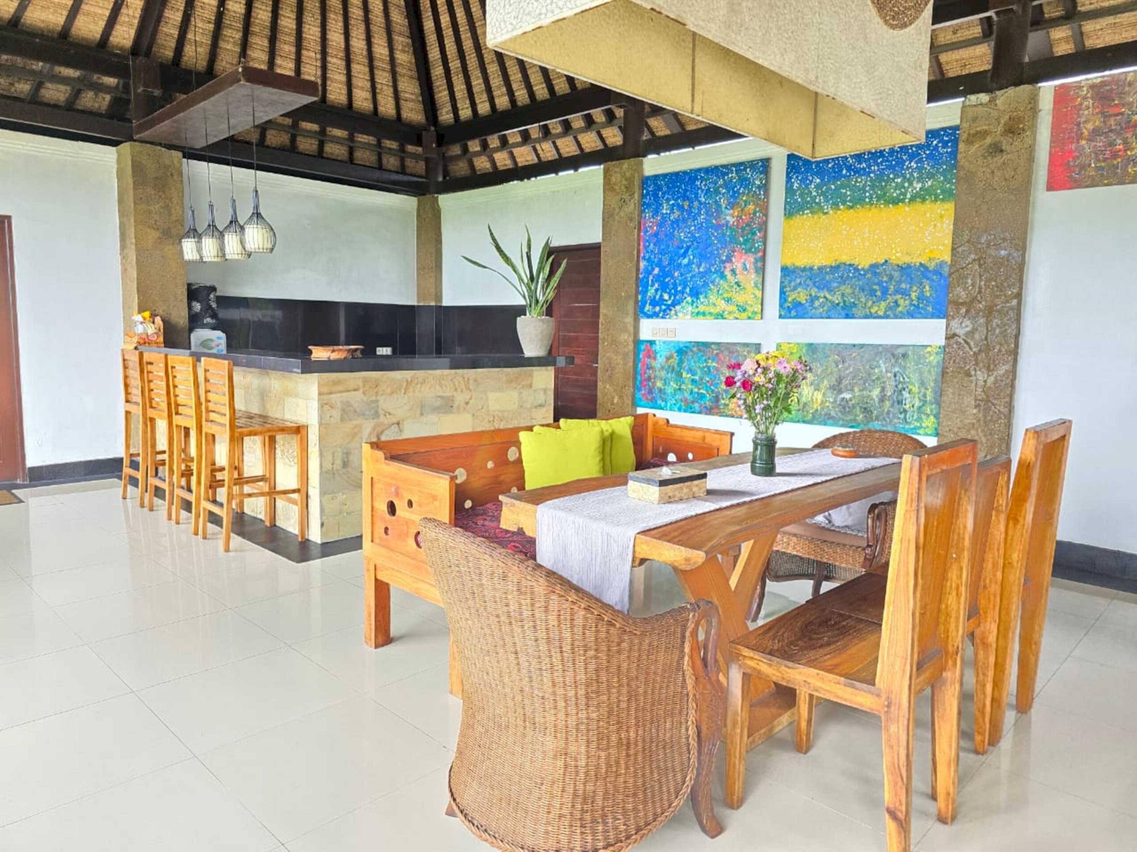 Un comedor amplio con una mesa y sillas de madera, una zona de cocina con encimera negra, y un ventanal con coloridas obras de arte.