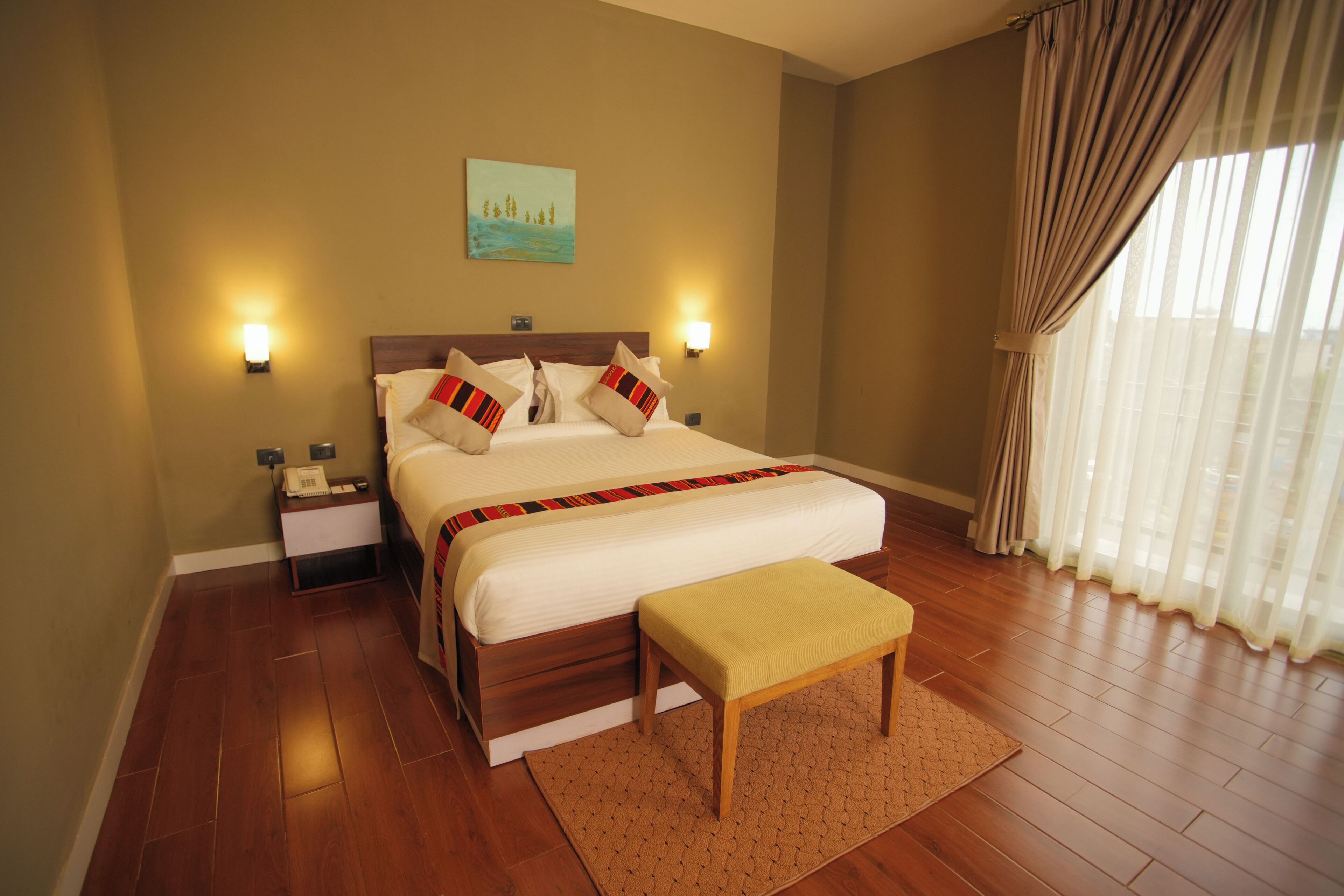 A hotel room with a large bed, bedside lamps, a nightstand with a phone, a wooden bench, and a painting on the wall.