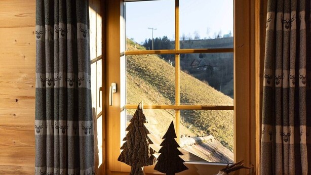 Een raam met uitzicht op een glooiend landschap, gordijnen met hertenprint en een kleine kerstboom op de vensterbank.