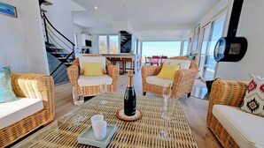 A spacious living room with wicker furniture, a bottle of champagne, and two glasses on a woven table. - Aux 3 Bains - Plage, Piscine, Spa (Locquirec)
