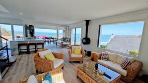 A spacious living area with wicker furniture, a kitchen, and large windows offering ocean views. - Aux 3 Bains - Plage, Piscine, Spa (Locquirec)