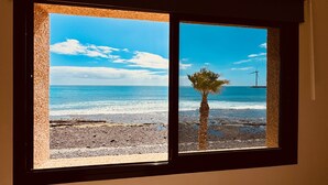 A window with a view of a beach, palm tree, and wind turbine. - Bright Sun House with Sea Views (Agüimes)
