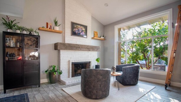 A modern living room with a fireplace, a dark cabinet, and a large window with a view of greenery. - Yorba Linda Furnished Retreat with Pool and Spa (Yorba Linda)