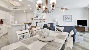 Une cuisine moderne avec des meubles blancs, un coin repas comprenant une table et des chaises blanches, un canapé gris et un ventilateur de plafond.
