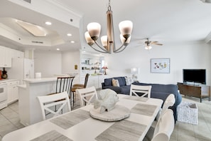 Une cuisine moderne avec des meubles blancs, un coin repas comprenant une table et des chaises blanches, un canapé gris et un ventilateur de plafond.