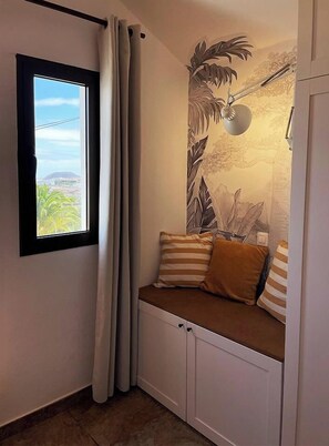 Une pièce d’angle avec une fenêtre, un banc agrémenté de coussins, une lampe fixée au mur et une vue partielle sur un paysage extérieur.