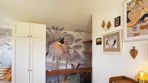 Ein kleines Zimmer mit einem Bett, einem Schreibtisch und einem Stuhl. Es gibt einen weißen Kleiderschrank, eine Wand mit einem tropischen Blattmuster und ein kleines Regal mit Dekoration.