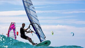 Una persona practica windsurf a la platja sota un cel clar i aigües tranquil·les.