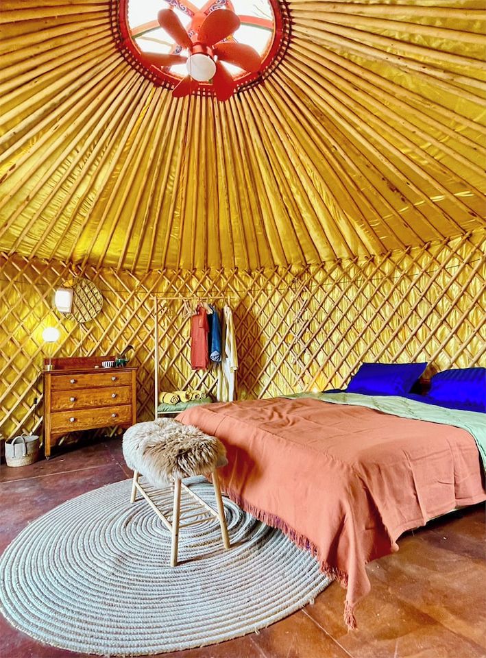 Una habitación con techo circular, cama con edredón rosa, cómoda de madera, alfombra redonda y lámpara colgante.