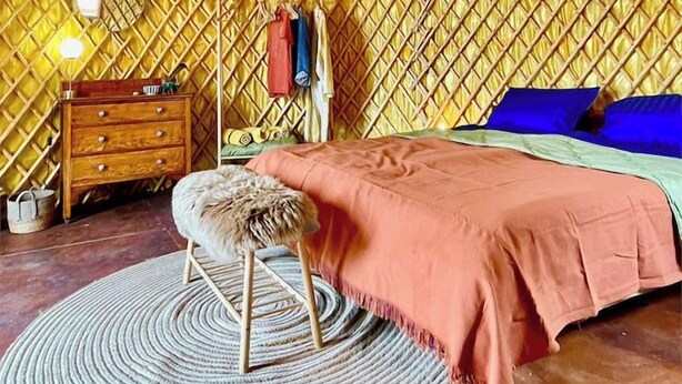 Una habitación con techo circular, cama con edredón rosa, cómoda de madera, alfombra redonda y lámpara colgante.