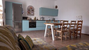 A compact kitchen with teal cabinets, a stainless steel refrigerator, and a dining area with a wooden table and chairs. - Co-Living at Casa Cocolores (La Oliva)