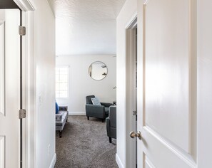 Un couloir mène à un espace de vie comprenant un canapé gris, un miroir rond accroché au mur et une porte dotée d’une poignée ronde.