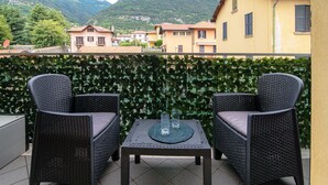 Una terraza con dos sillas de mimbre y una mesita, con vistas a una montaña y una hilera de edificios amarillos.