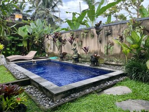 A rectangular swimming pool with a stone border, surrounded by tropical plants and a concrete wall. - Belvilla Abhinaya Central Ubud (Payangan)