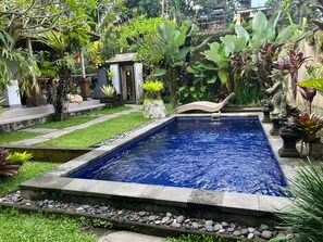 A rectangular swimming pool with clear blue water, surrounded by a stone border and a grassy area with various plants. - Belvilla Abhinaya Central Ubud (Payangan)
