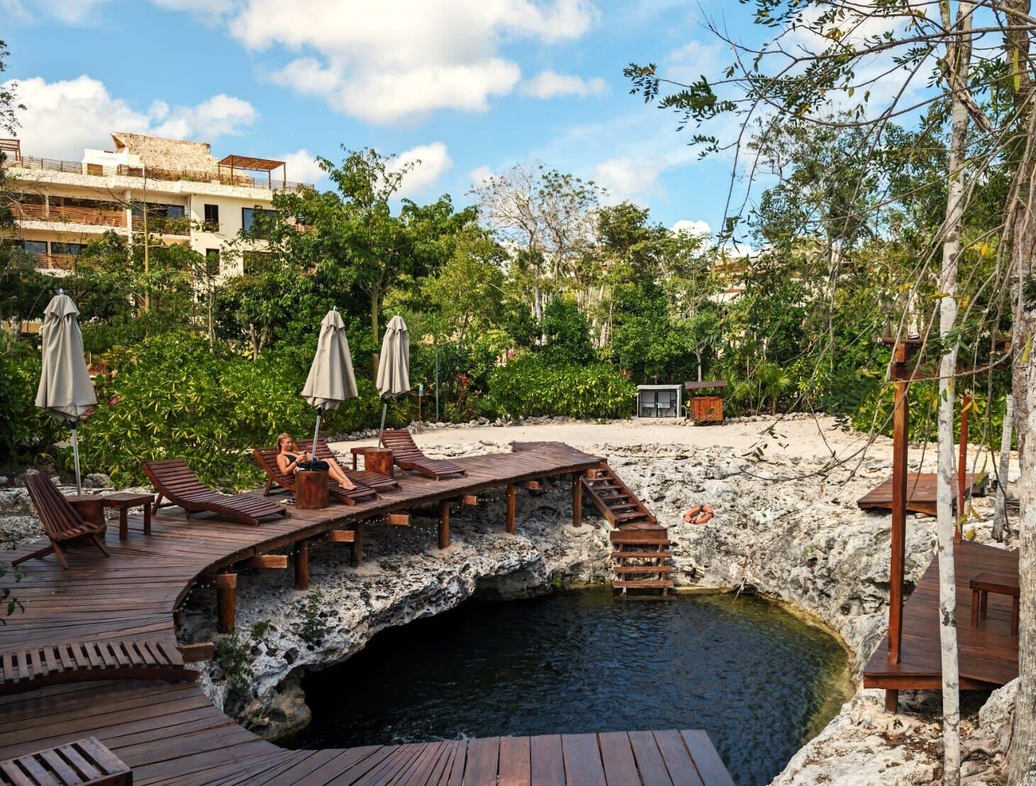 A wooden deck with lounge chairs and umbrellas overlooking a natural pool surrounded by greenery and a rocky landscape.