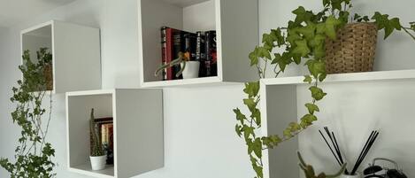Uma parede com várias prateleiras brancas, algumas com livros e plantas, e um relógio na prateleira de baixo.
