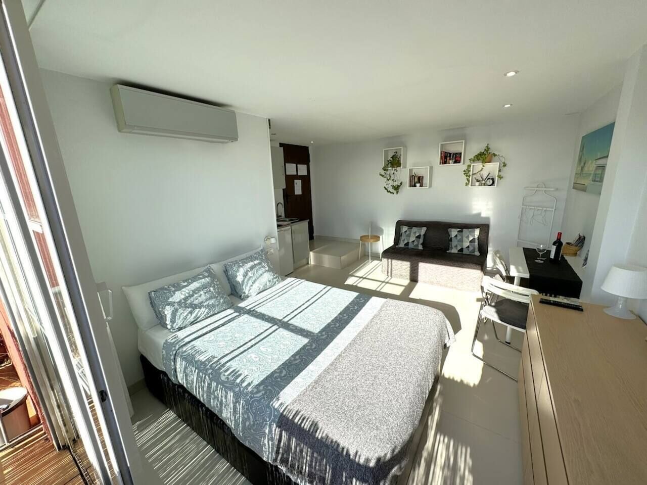 A modern bedroom with a large bed, a desk, and a chair. There is a small table with a lamp and a picture frame.