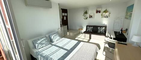 A modern bedroom with a large bed, a desk, and a chair. There is a small table with a lamp and a picture frame.