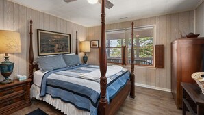 Un lit à baldaquin avec une courtepointe, une table de chevet avec une lampe, un tableau au mur et une fenêtre avec des volets.