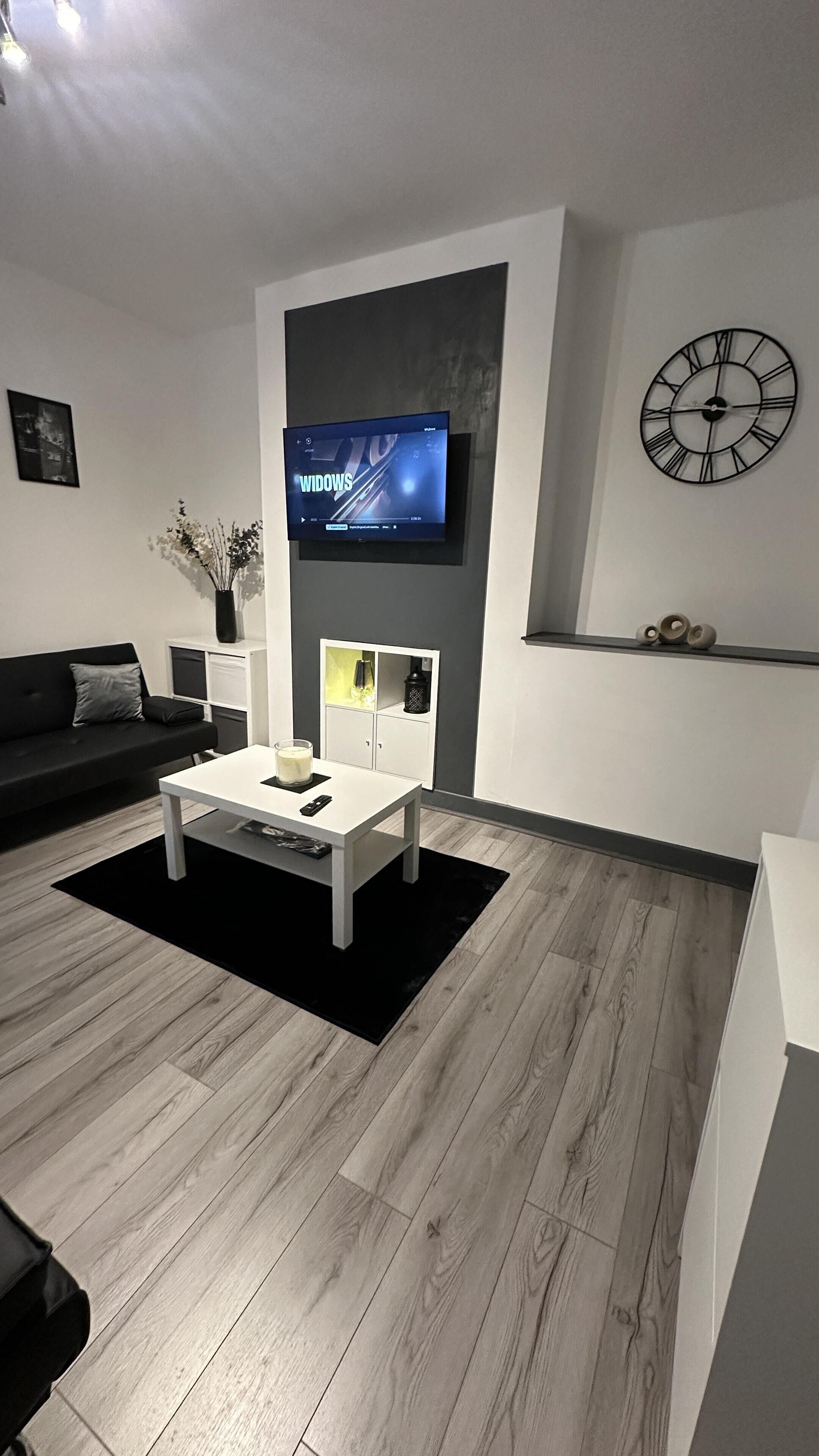 A modern living room with a flat-screen TV mounted on a wall, a white coffee table, a black sofa, and a wooden floor.