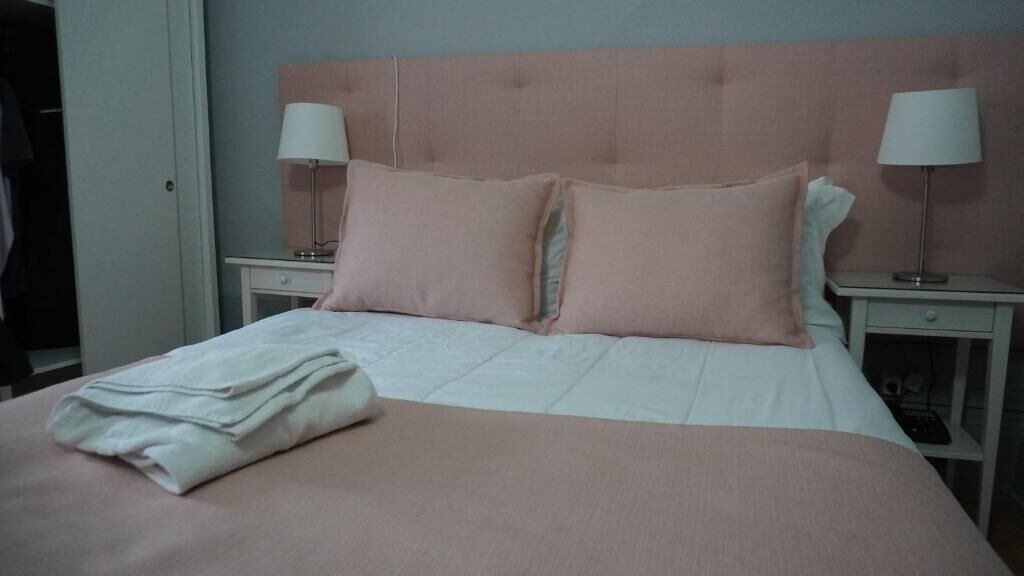 Un lit bien fait, avec une tête de lit rose, deux tables de chevet avec des lampes et une serviette blanche pliée posée sur le lit.