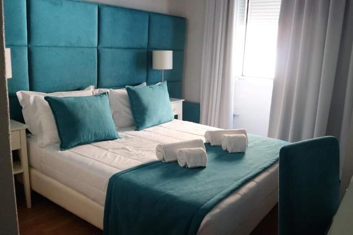 Une chambre d’hôtel avec un grand lit, une tête de lit turquoise, du linge de lit blanc et une fenêtre avec des rideaux.