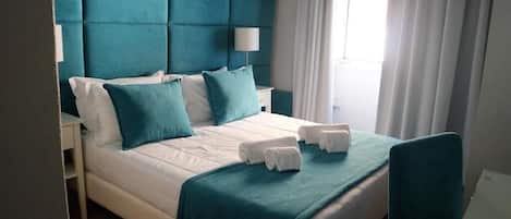 A hotel room with a large bed, teal headboard, white bedding, and a window with curtains.