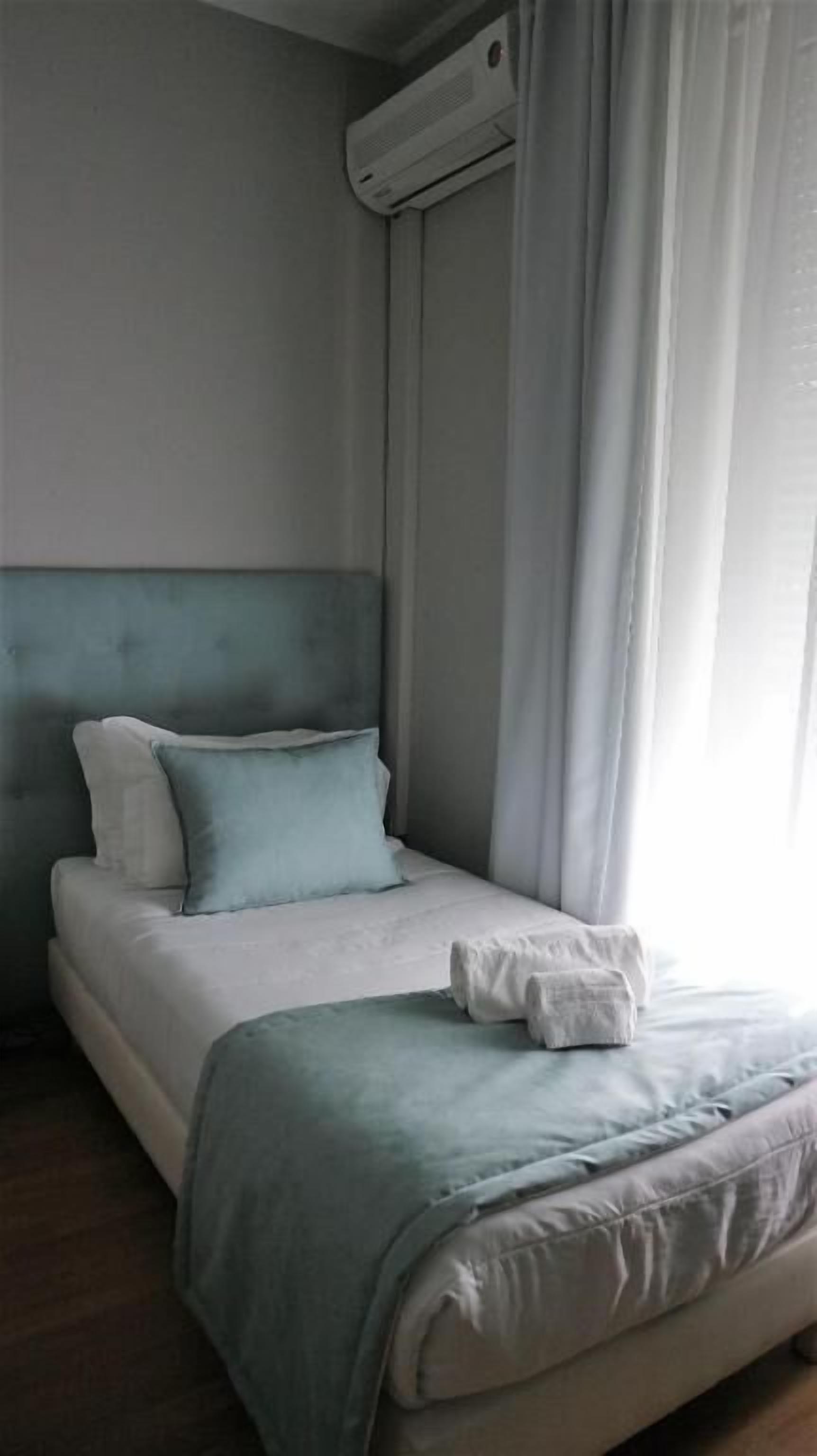 Una cama bien hecha con cabecera azul, almohadas blancas y manta azul. Se observa un equipo de aire acondicionado montado en la pared sobre la cama.