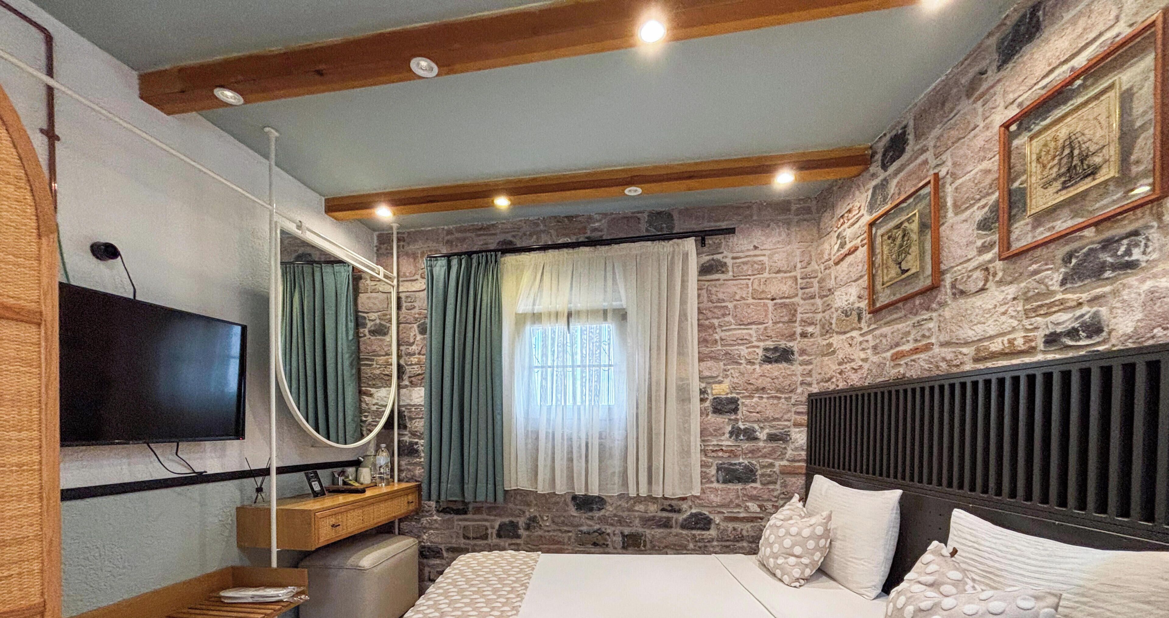 Un dormitorio con pared de piedra, cama con ropa de cama blanca, cabecero de madera, televisor de pantalla plana, tocador con espejo y ventana con cortinas.