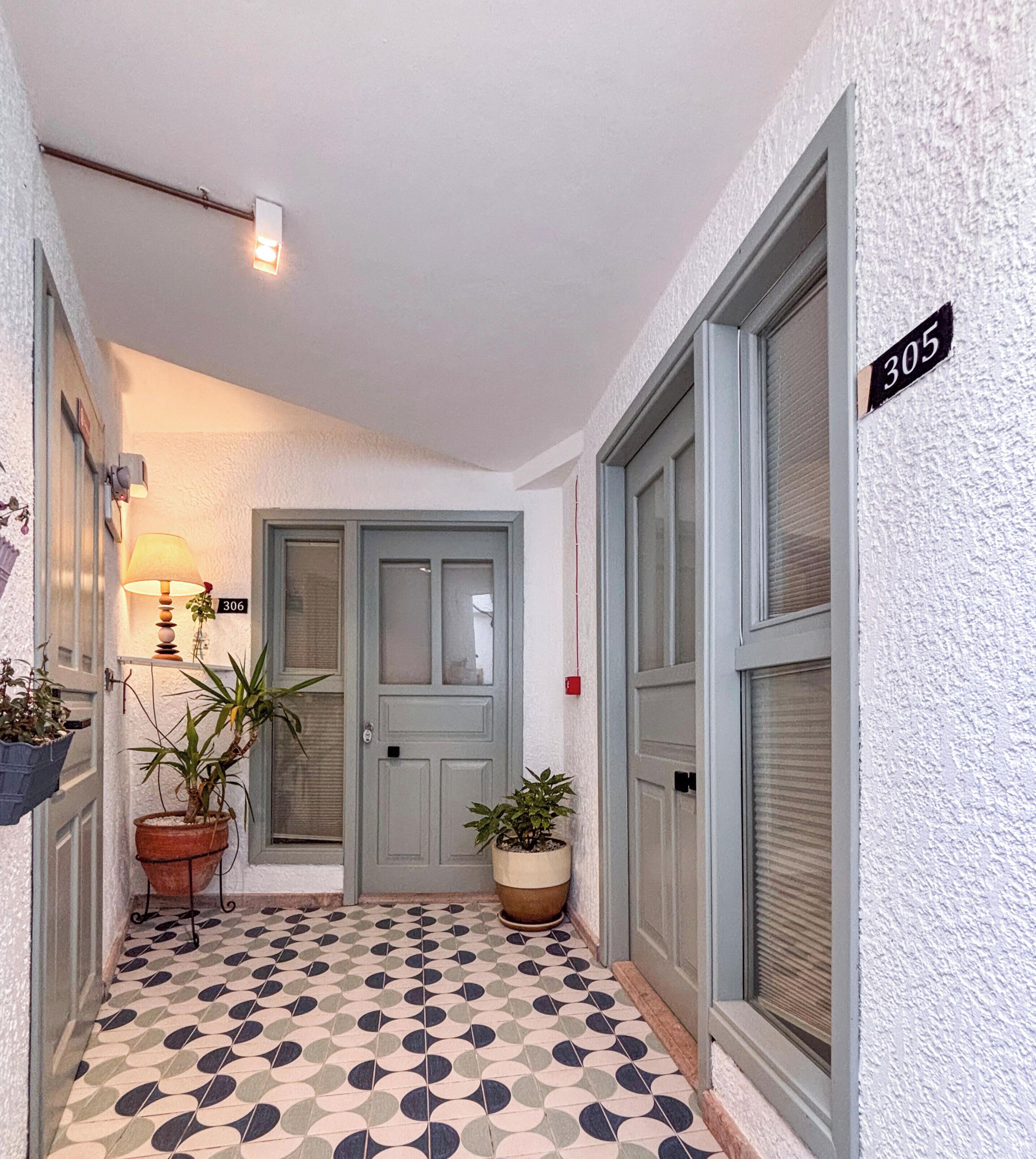 Un pasillo con piso estampado, una puerta con placa de número y plantas en macetas.