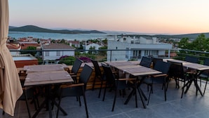 An outdoor terrace with tables and chairs, overlooking a body of water and a town. - Qara Otel (Ayvalik)