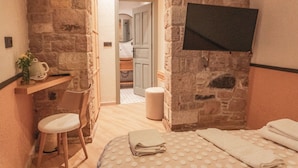 A cozy room with a stone wall, a wooden chair, a bed with a polka dot blanket, and a flat-screen TV mounted on the wall. - Qara Otel (Ayvalik)
