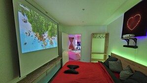 Une chambre avec une grande télévision, un lit avec une literie rouge, un miroir et un projecteur posé sur une étagère.