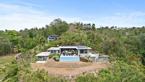A modern house with a pool, surrounded by dense vegetation and other houses on a hillside.
